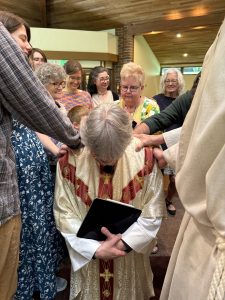Vicar James, a gray-haired white man, kneels while members of the church gather around and lay hands on him to bless him.